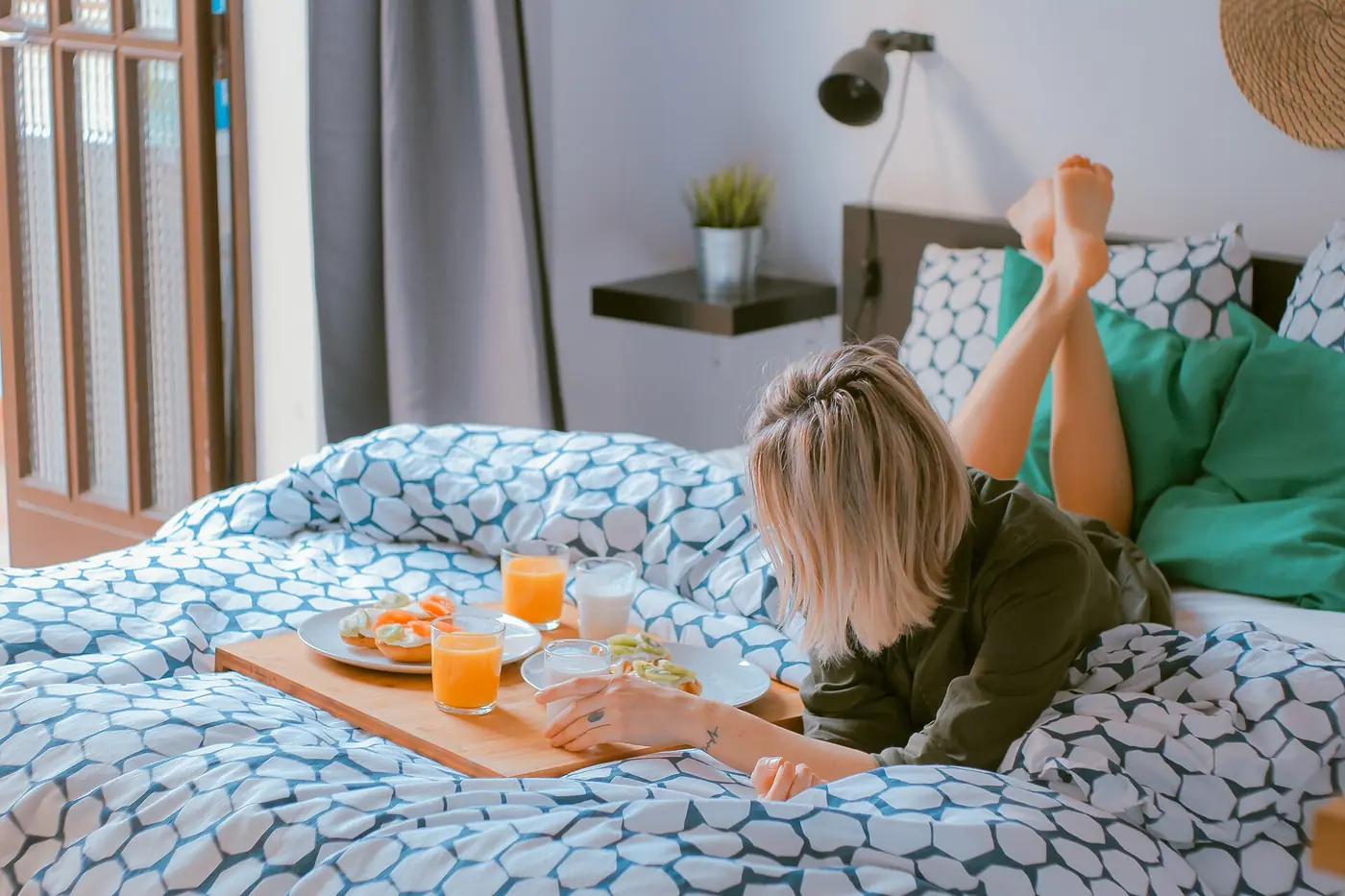 Mujer lee recostada en la cama junto a una charola con jugo y pan